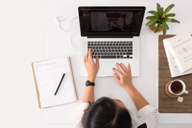 Business person sits at a table and works on a laptop with notebook and cup of coffee nearby