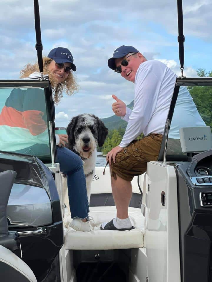 Two people with large dog smile and give thumbs up from boat deck